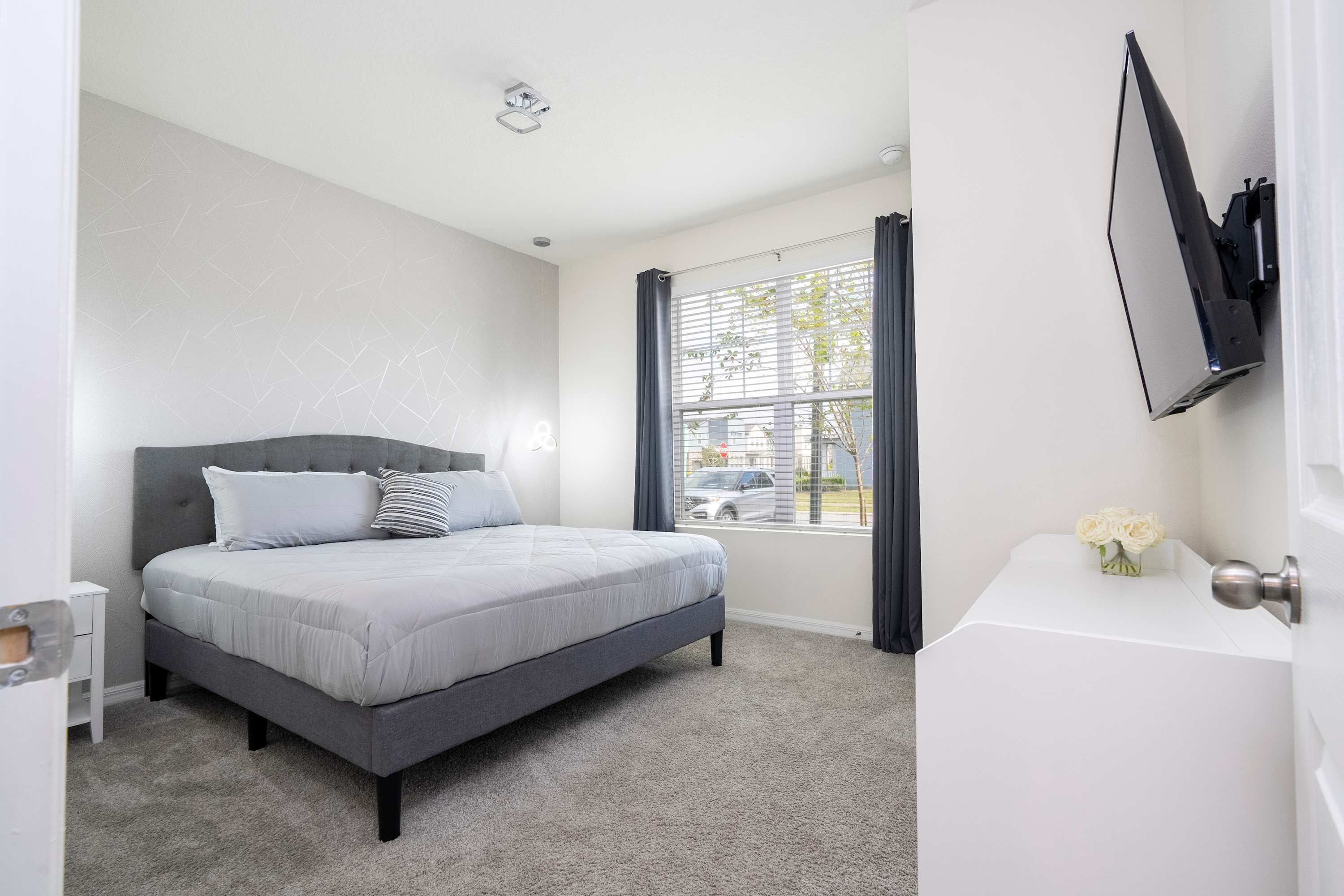 A modern bedroom with a bed, a bedside table, a window with curtains, and a flat-screen TV mounted on the wall.