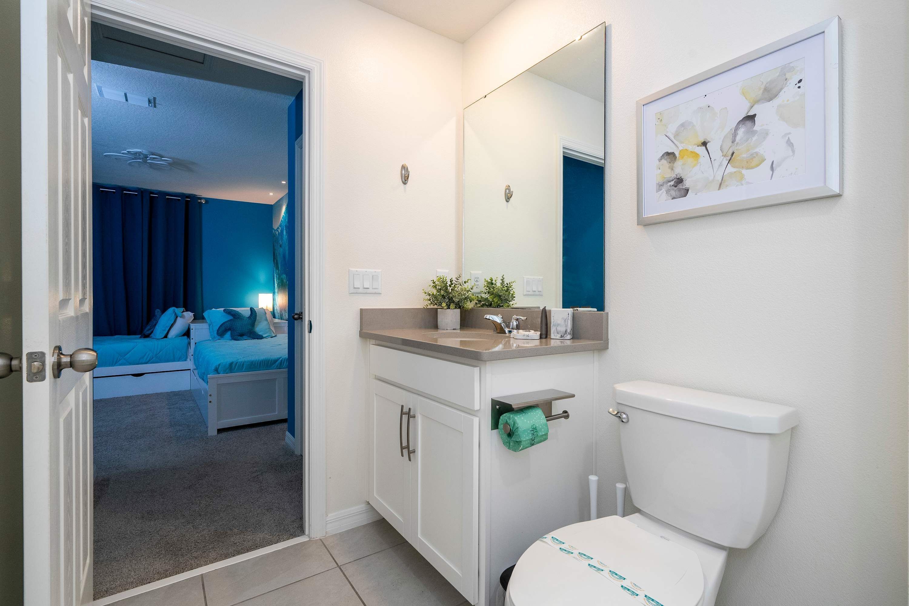 Une salle de bain avec des toilettes, un lavabo et un miroir. Un tableau encadré est accroché au mur et une plante est posée sur le comptoir.