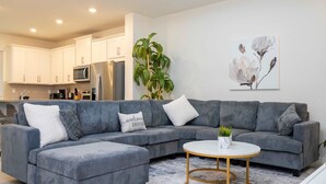 A modern living room with a grey sectional sofa, a round coffee table, and a kitchen in the background. - Family Fun Moana Mario Harry Potter Stay (Kissimmee)