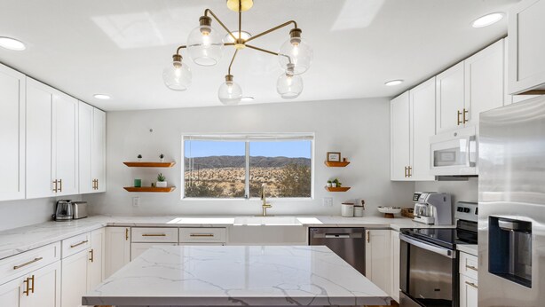 A modern kitchen with white cabinetry, a marble island, stainless steel appliances, and a view of a desert landscape through a large window. - Private compound, offering three separate living spaces! multi families,groups (Desert Hot Springs)