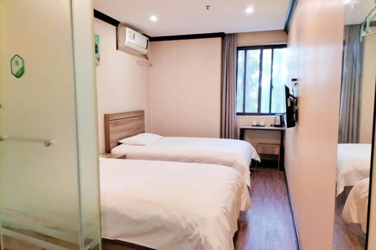 A hotel room with two beds, a small desk, a window with curtains, and an air conditioning unit.