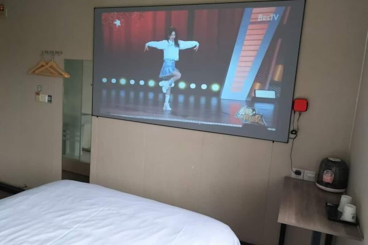 A hotel room with a bed, a TV mounted on the wall displaying a performance, a small table with a kettle and cups, and a wooden hanger on the wall.