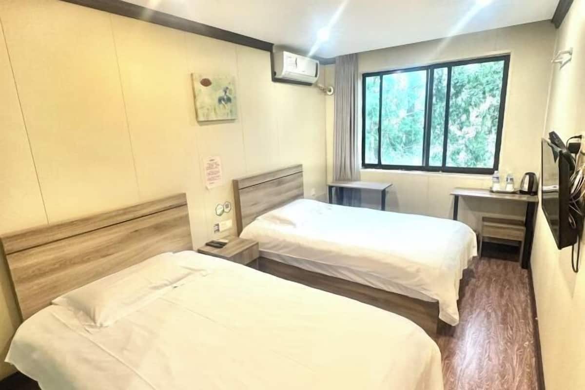 A hotel room with two beds, a desk, a chair, a window with a view, and a wall-mounted air conditioner.
