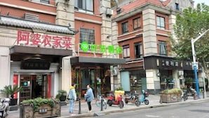 A street view with a building featuring a traditional Chinese architectural style, a sign for a convenience store, and a few pedestrians and scooters on the sidewalk.