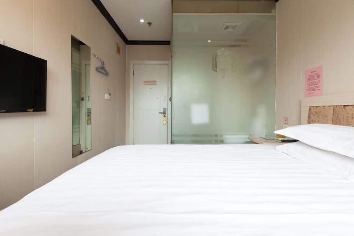 A hotel room with a bed, a television mounted on the wall, a glass shower enclosure, and a door with a sign.