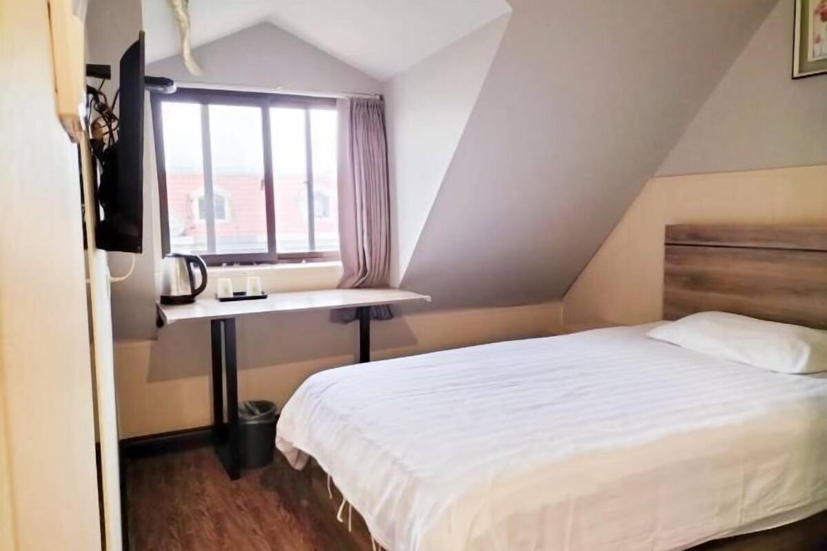 A bedroom with a bed, a window, a table with a kettle, and a wall-mounted TV.