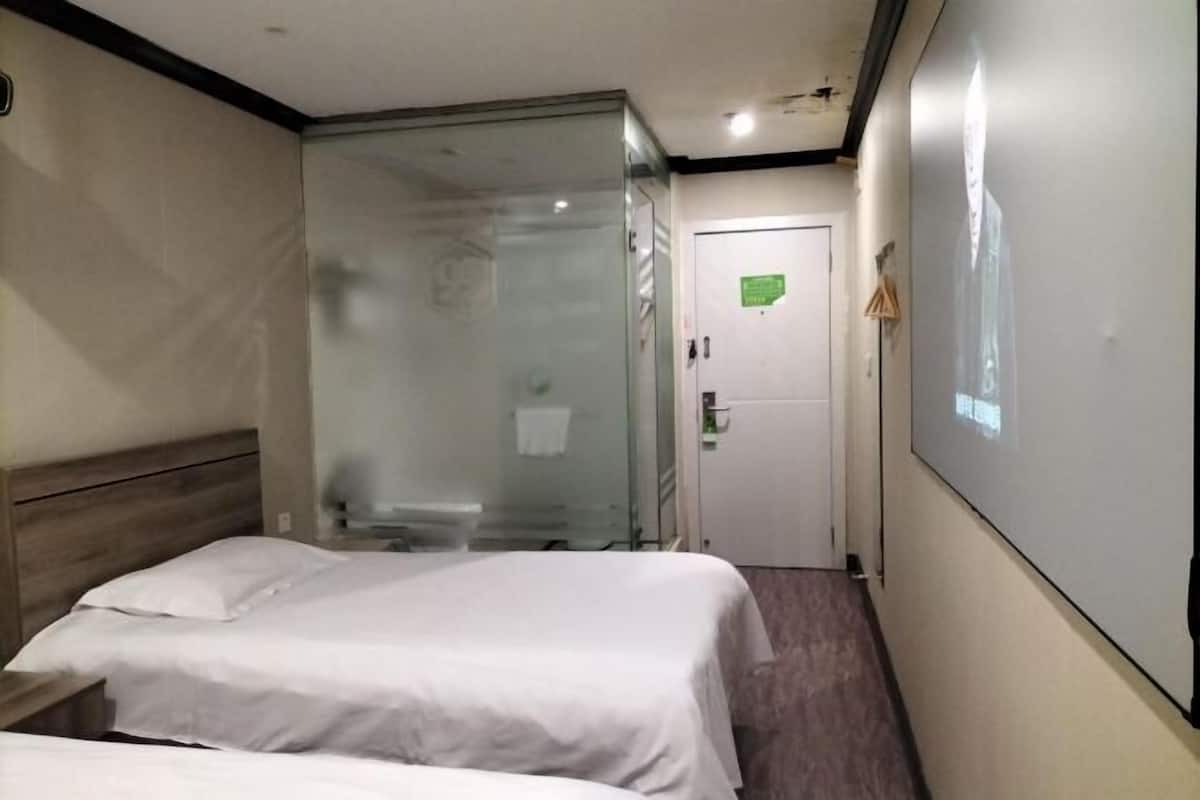 A hotel room with two beds, a glass shower enclosure, a white door, and a projection screen on the wall.