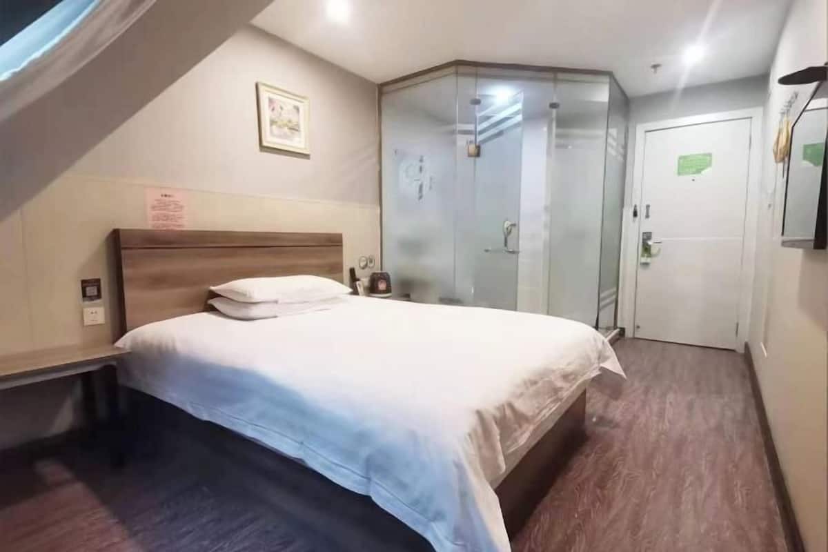A hotel room with a bed, a bedside table, a nightstand, a glass-enclosed shower, and a door.