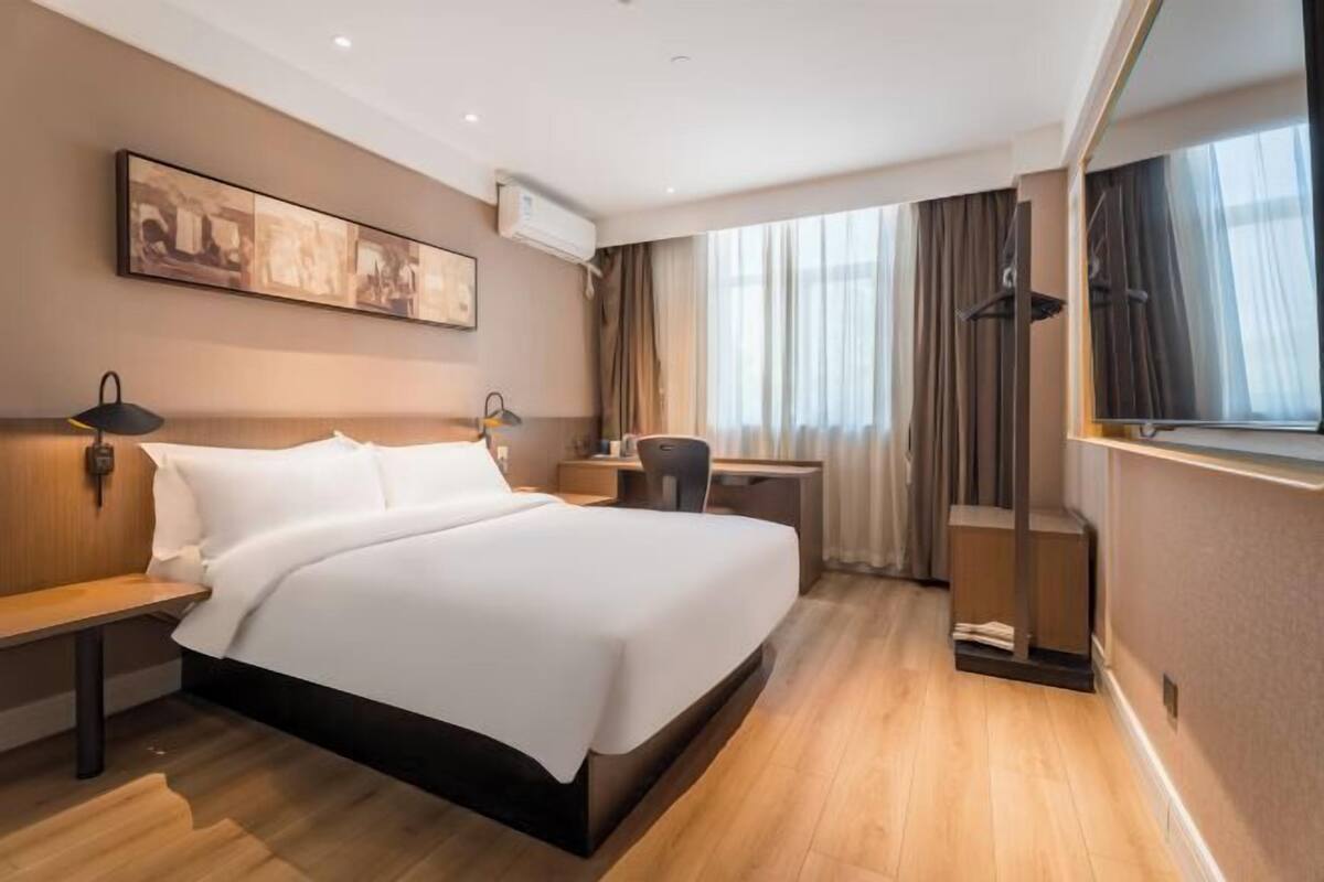 A hotel room with a bed, bedside tables, a desk, a chair, a hanging light, a window with curtains, and a wall-mounted air conditioner.