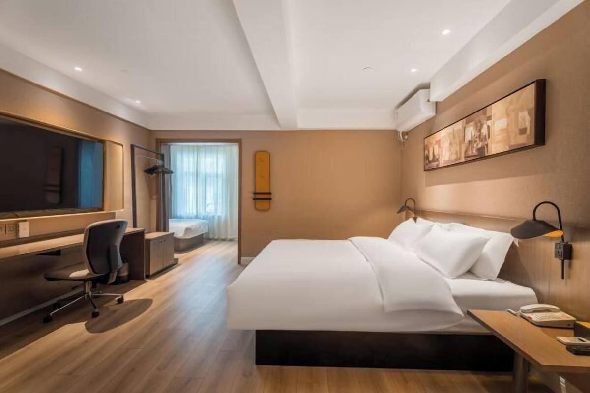 A modern hotel room with a large bed, a desk with a chair, a desk lamp, a telephone, and a painting on the wall.