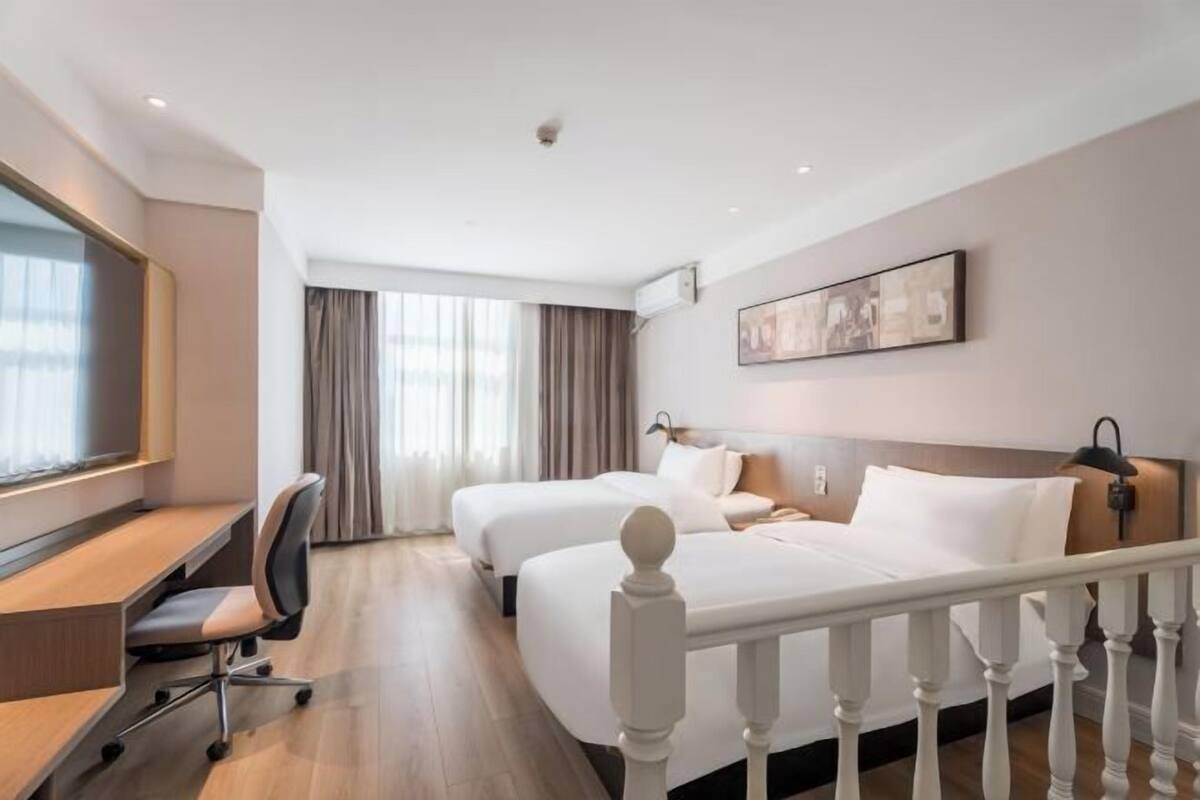 A hotel room with two beds, a desk, a chair, a window with curtains, and a painting on the wall.