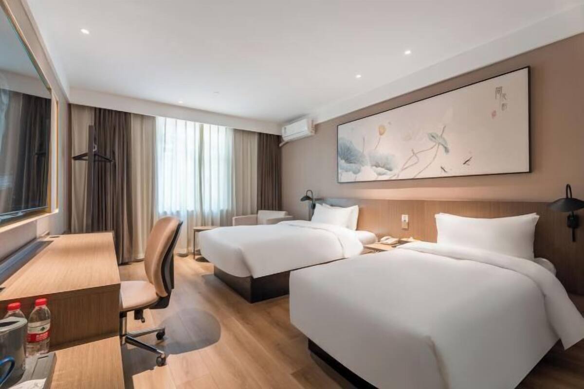 A hotel room with two beds, a desk, a chair, a large abstract painting, and a window with curtains.