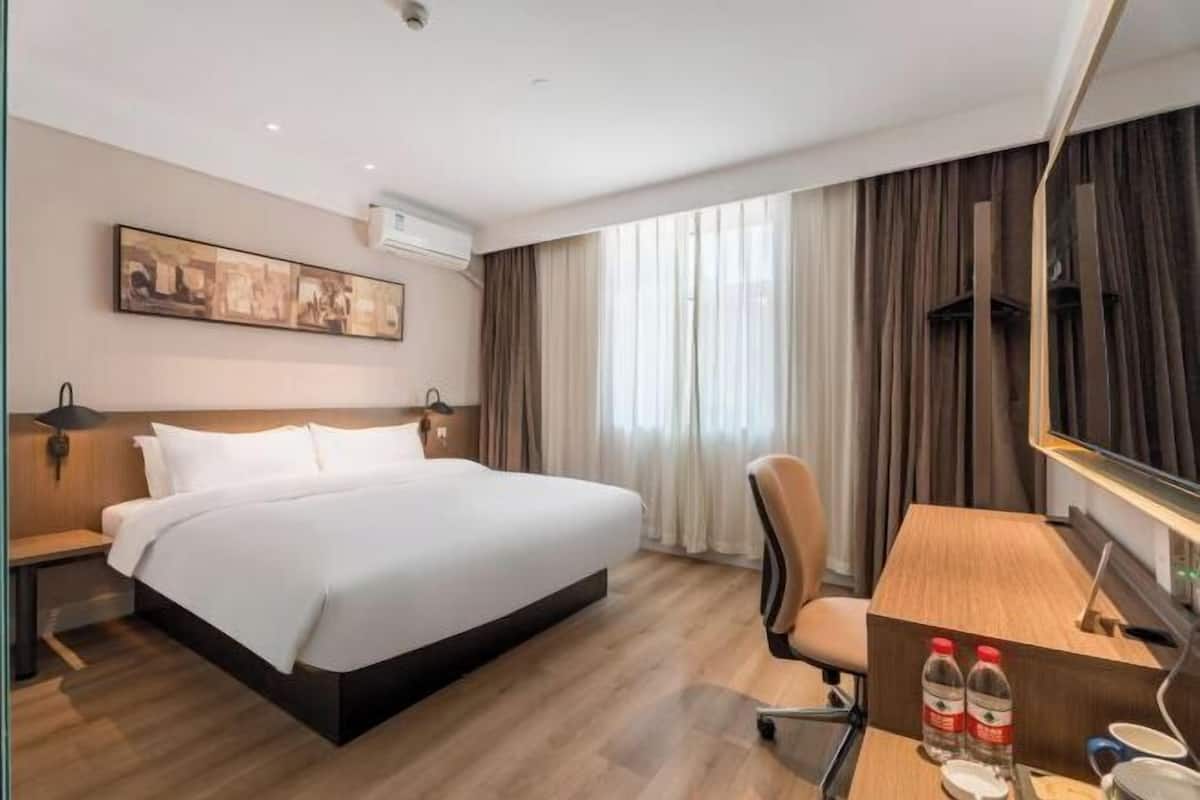 A hotel room with a large bed, a desk, a chair, a painting on the wall, and a window with curtains.