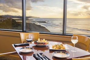 A dining table with plates of food, wine glasses, and a view of the ocean through large windows. - Inn at Spanish Head (Lincoln City)