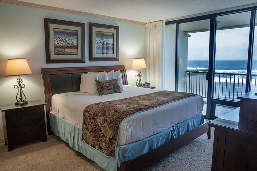 A hotel room with a large bed, two bedside lamps, a nightstand, a desk, and a balcony with a view of the ocean.