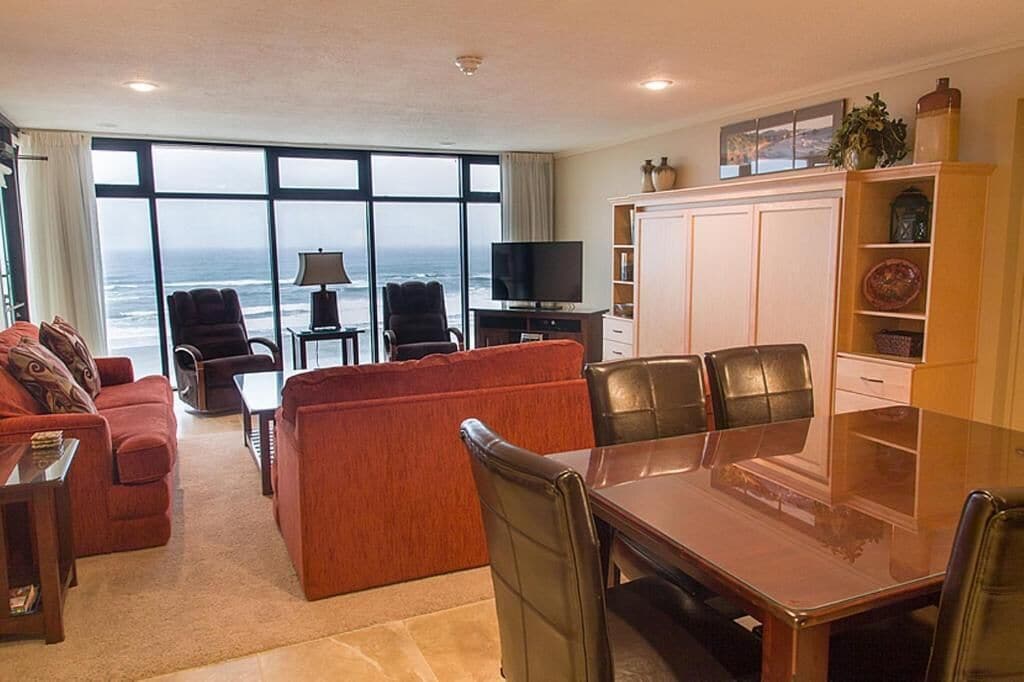 A spacious living room with a large window overlooking the ocean, a dining area with a table and chairs, and a television on a stand.