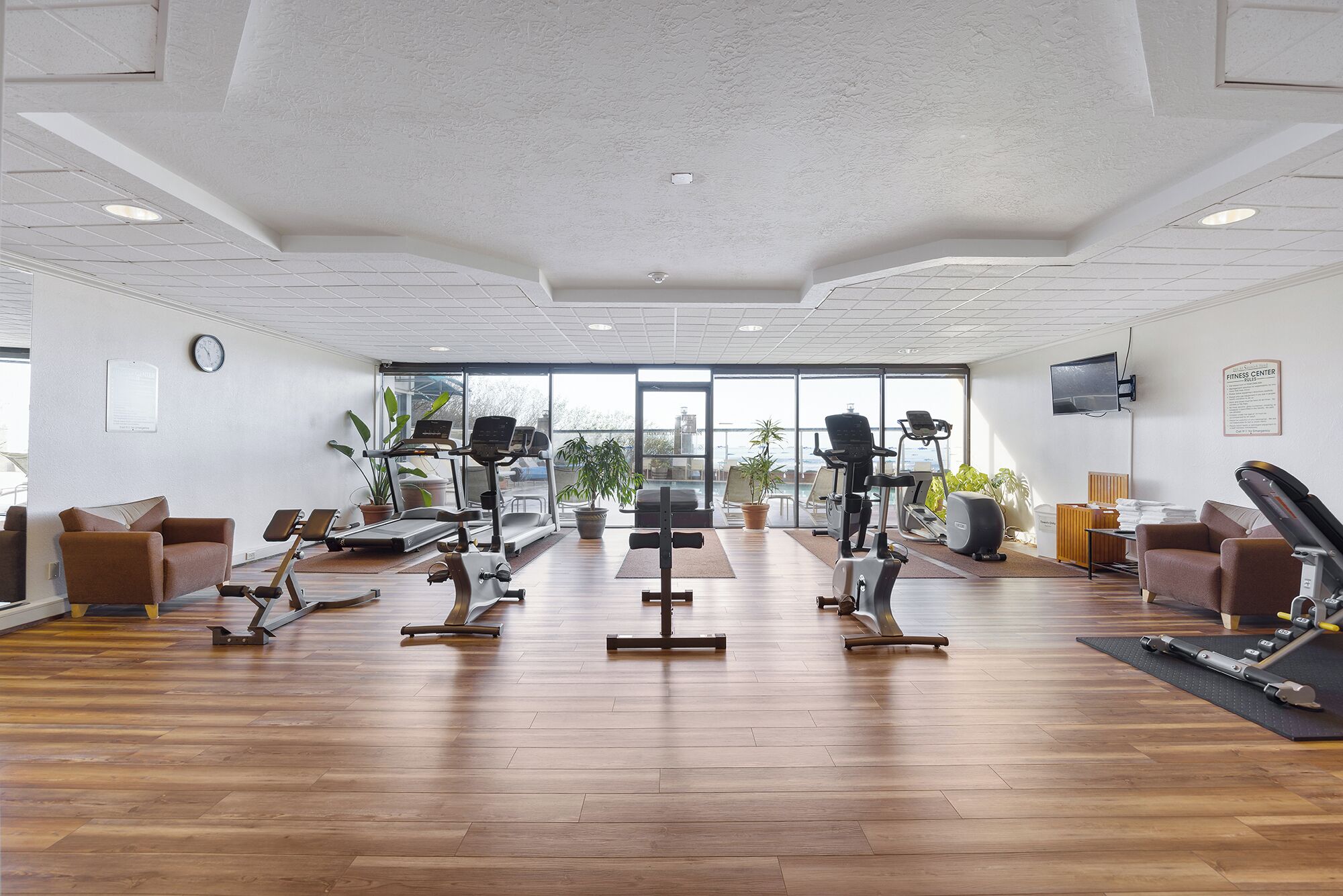 Un gimnasio amplio con cintas de correr, bicicletas estáticas y máquinas de pesas, ventanas grandes y un reloj en la pared.