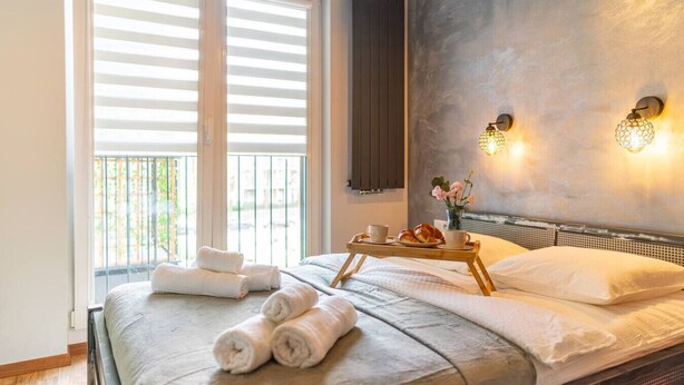 A hotel room with a bed, towels, a tray with croissants and coffee, and a view of the outdoors through large windows.