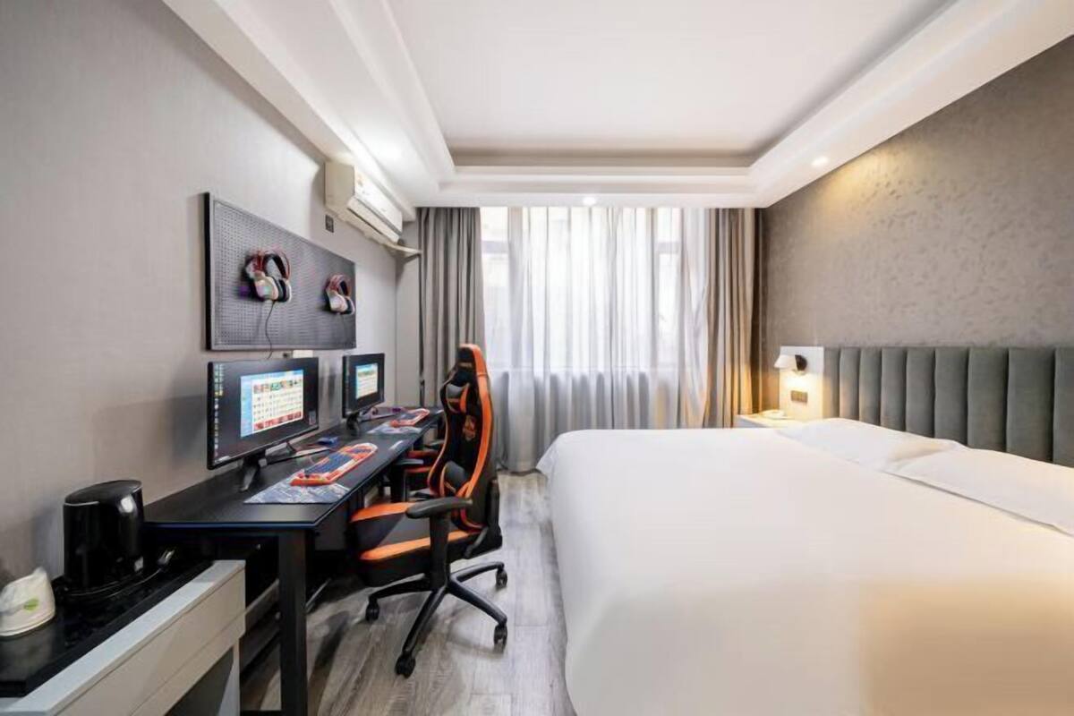 A hotel room with a bed, two monitors, a desk, an office chair, a coffee maker, and a window with curtains.