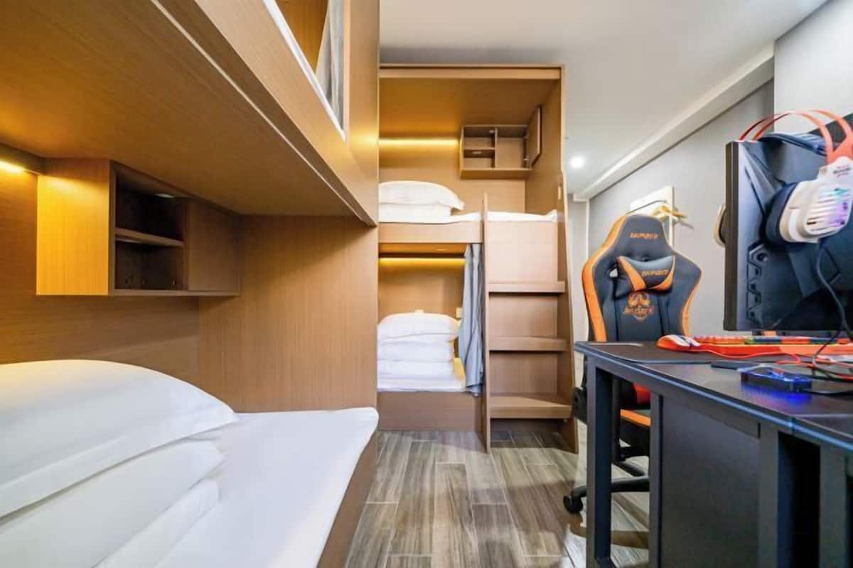 A compact hotel room with a bunk bed, a desk with a computer, and a gaming chair.