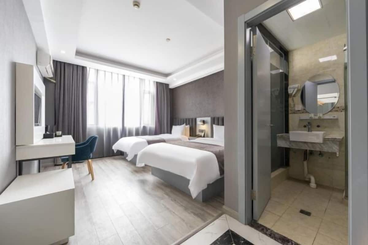A hotel room with a desk, two beds, a bathroom with a sink and mirror, and a view of the outdoors.