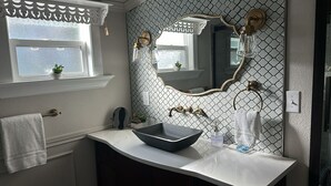 Une salle de bain avec un lavabo blanc, un grand miroir et une fenêtre agrémentée d’une lambrequin blanche.
