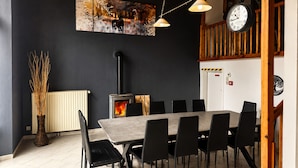 A dining area with a long table, chairs, a wood-burning stove, a clock, and a wall-mounted painting.
