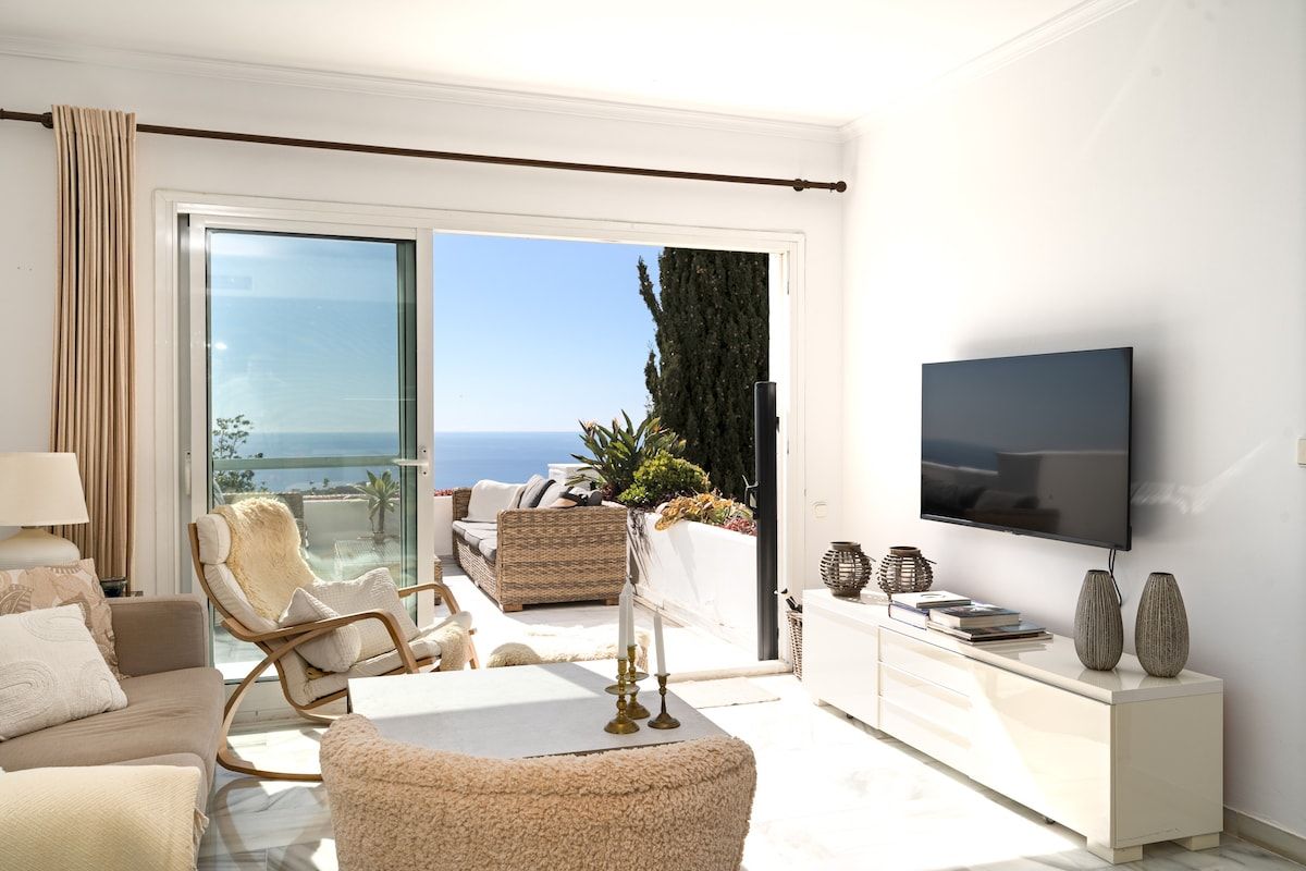 A modern living room with a large TV, comfortable seating, a balcony with a view, and a white interior.