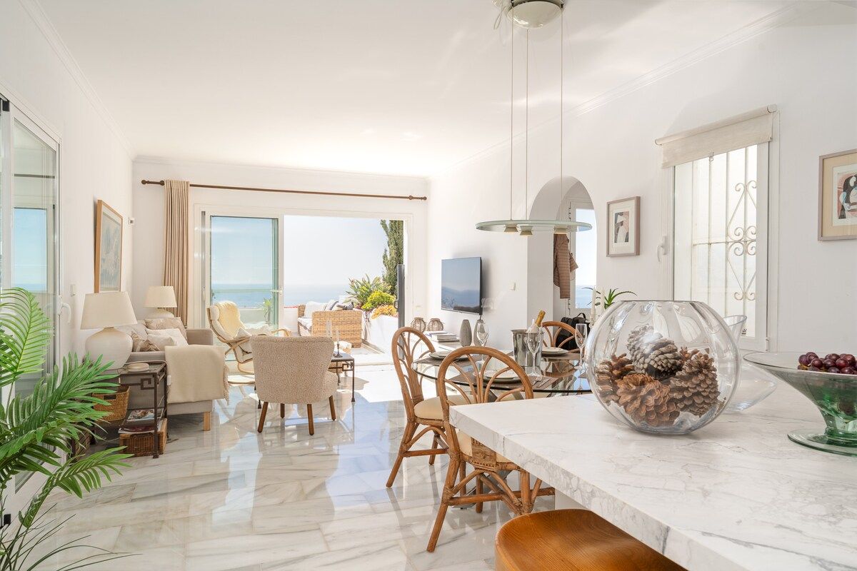 A spacious living area with a view of the sea, a dining table with chairs, and a kitchen area with a TV.