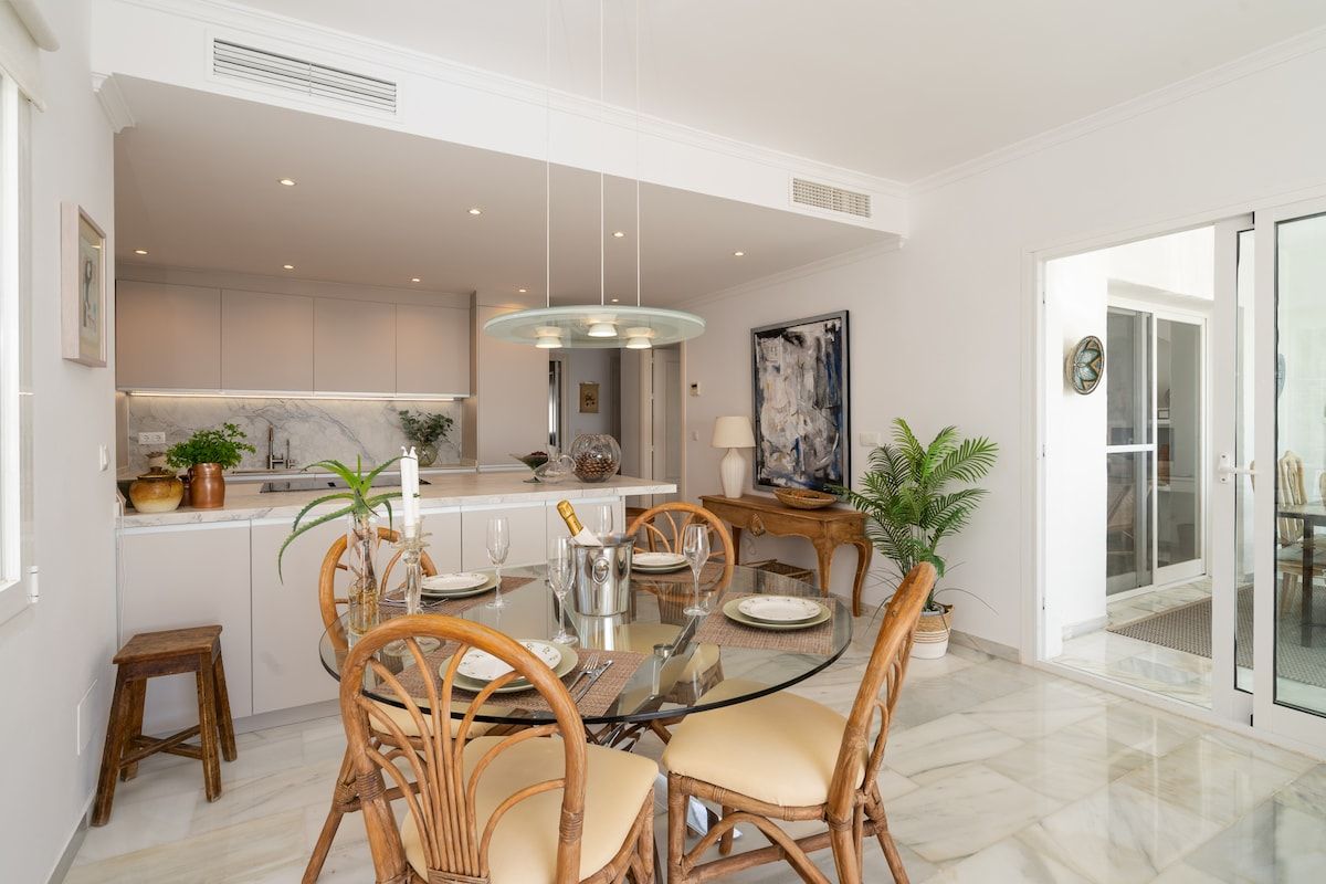 A modern dining area with a glass table, wicker chairs, and a kitchen in the background.