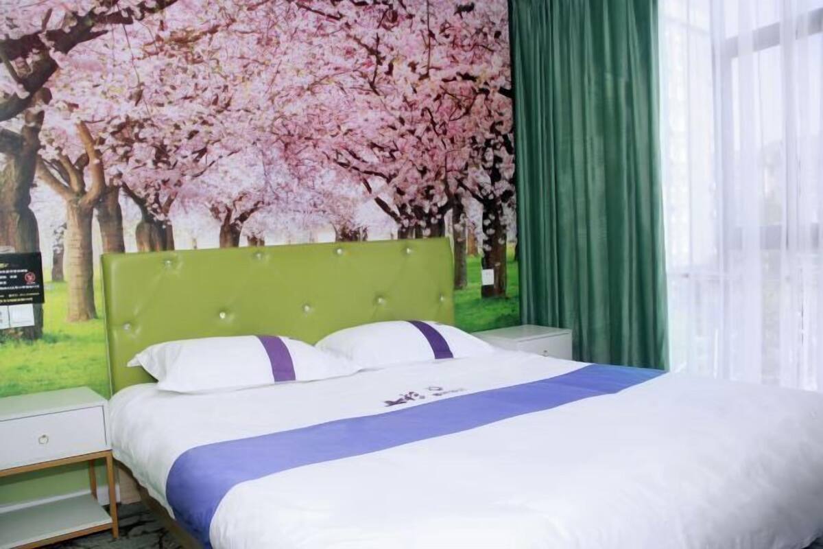 Ein Hotelzimmer mit einem grünen Kopfteil, Bettwäsche in Weiß und Lila sowie einer Kirschblüten-Wandmalerei.
