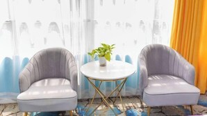 Una habitación con dos sillones grises, una pequeña mesa blanca con una planta y una ventana con cortina.