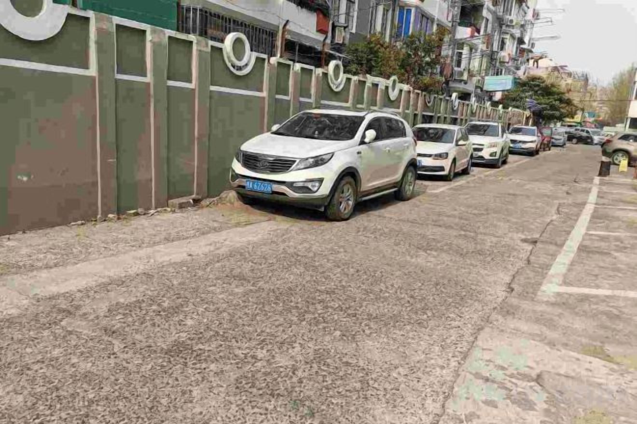 A row of parked cars along a street with a green fence on one side.