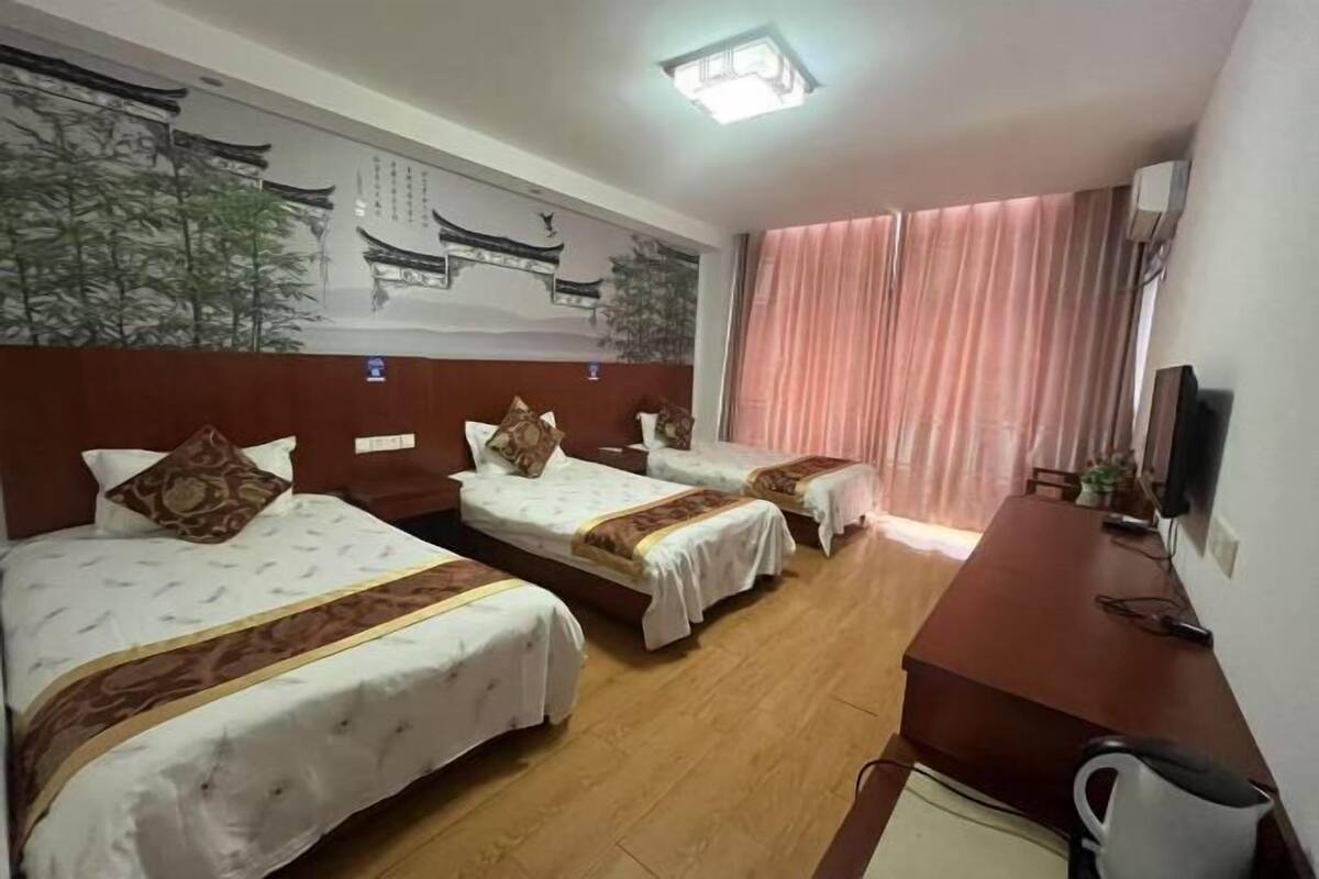 A hotel room with three beds, a wooden desk, a television, and a large window with curtains.