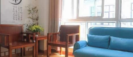 Une pièce avec un canapé bleu, des chaises en bois, une petite table avec une plante et une fenêtre avec des rideaux.