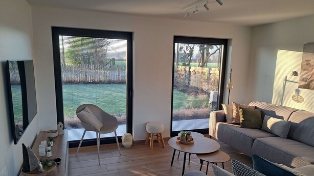 A modern living room with a grey sofa, a round coffee table, and a chair. There are large windows with blinds, a TV, and a small table with decorative items.