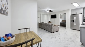 A modern living room with a sofa, a dining table with chairs, a TV mounted on the wall, and a kitchen area with a refrigerator.
