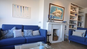 A living room with a blue sofa, a glass coffee table, and a fireplace with a framed shark painting above it. - Apartment in Belgium Near Seafront & Casino (Knokke-Heist)