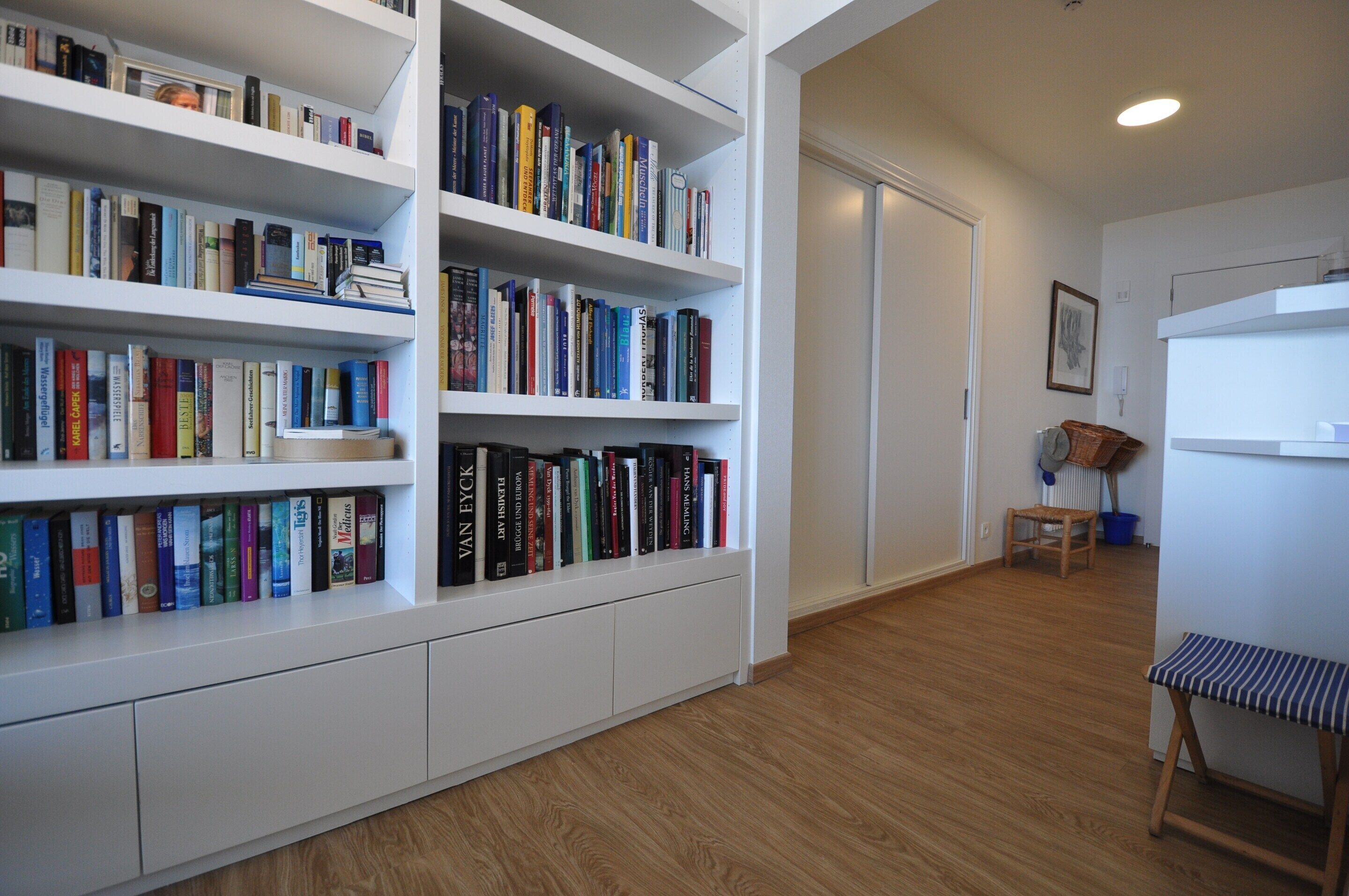 A room with white built-in bookshelves filled with books, a wooden floor, a small wooden bench, and a radiator.