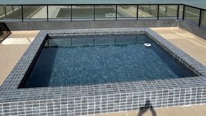 Piscina rectangular con bordes azulejados, rodeada por una barandilla de vidrio, con vistas al mar tranquilo bajo un cielo parcialmente nublado.