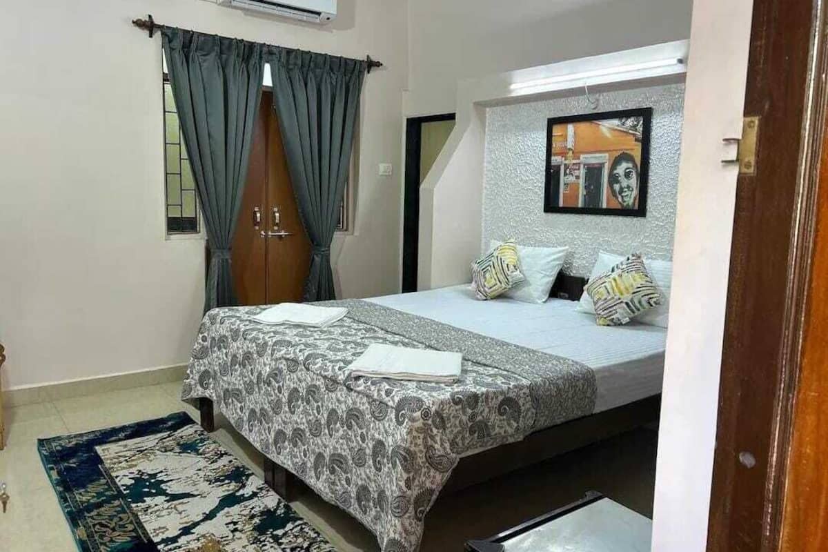 A bedroom with a bed, a window with curtains, a wall-mounted air conditioner, and a framed picture.