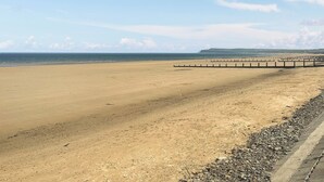 Una amplia playa arenosa con un muelle de madera que se adentra en el mar, cielo despejado y colinas al fondo.