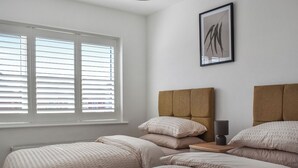 Un dormitorio con dos camas, una ventana con persianas, una mesita de noche con una lámpara y un cuadro enmarcado en la pared.