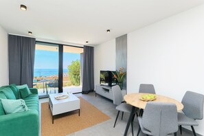 A modern living room with a sofa, coffee table, dining table, and chairs. There is a TV and a plant on a cabinet. The room has large windows with a view of the sea. - Hedera Estate, Hedera A60 (Dubrovnik)