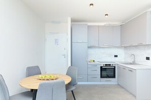 Una cocina moderna con armarios grises, horno integrado y un lavamanos. Hay una mesa redonda de madera con sillas y una cesta de manzanas.