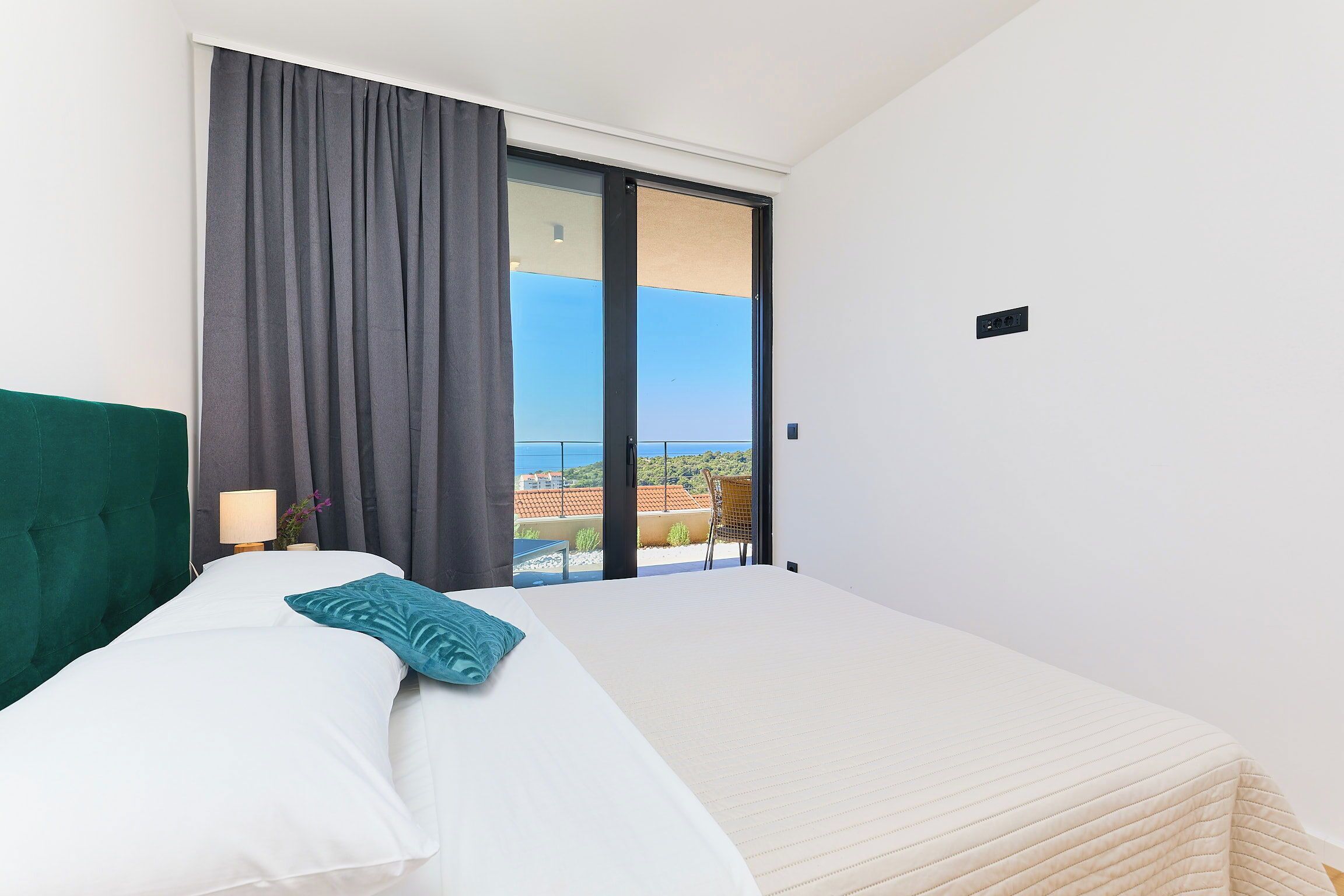 A bedroom with a bed, a nightstand, a lamp, and a view of a balcony.