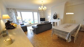 A modern living room with a sofa, a dining table, and a TV. There is a chandelier, a fireplace, and a view through the glass door. - Apartment in Duinbergen Near Sandy Beach (Knokke-Heist)