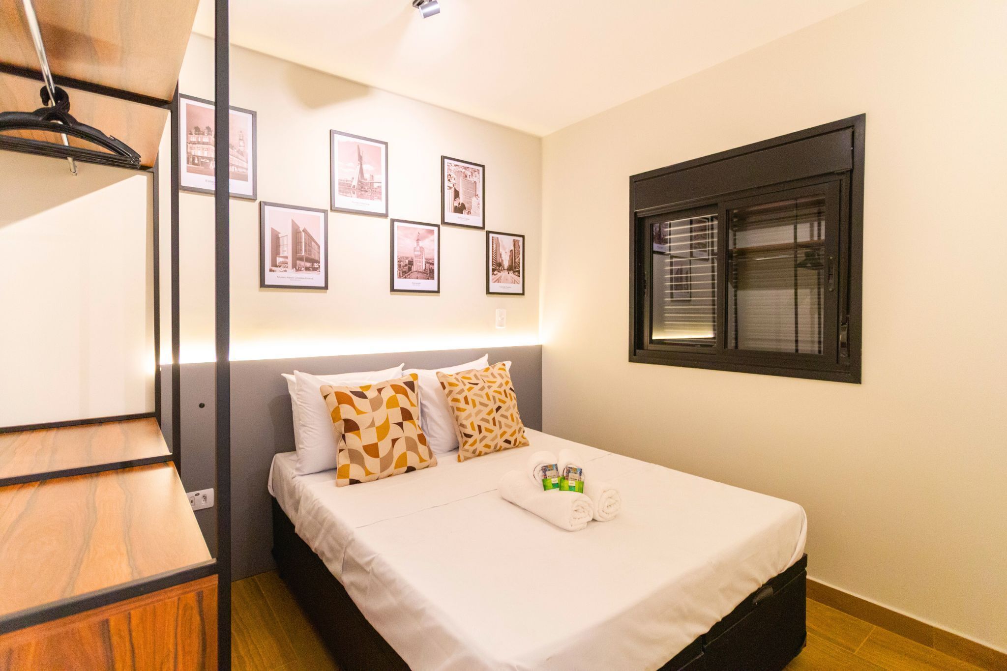 A hotel room with a bed, a wardrobe, a window with blinds, and several framed pictures on the wall.
