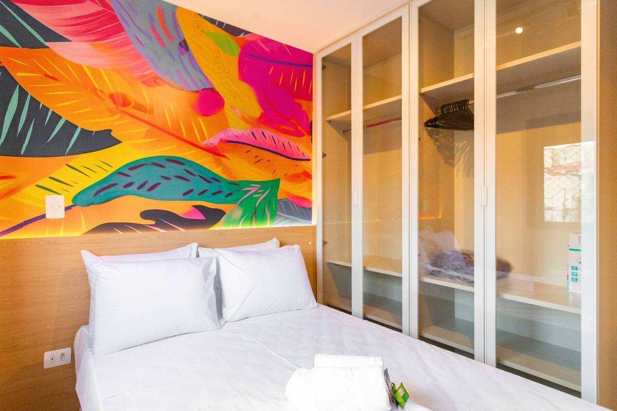 A hotel room with a bed, white linens, a colorful abstract mural on the wall, and a walk-in closet with glass doors.