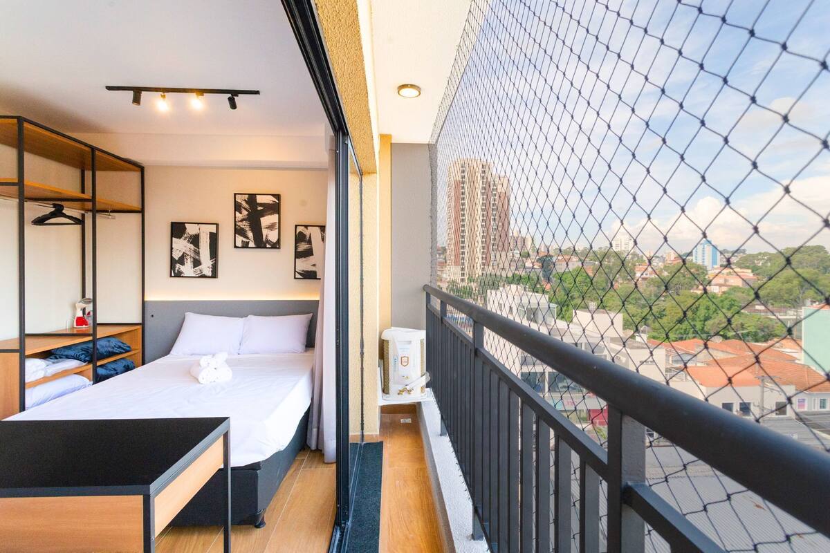 A balcony with a view of a cityscape, a bed with white linens, a small table, and a hanging chair.